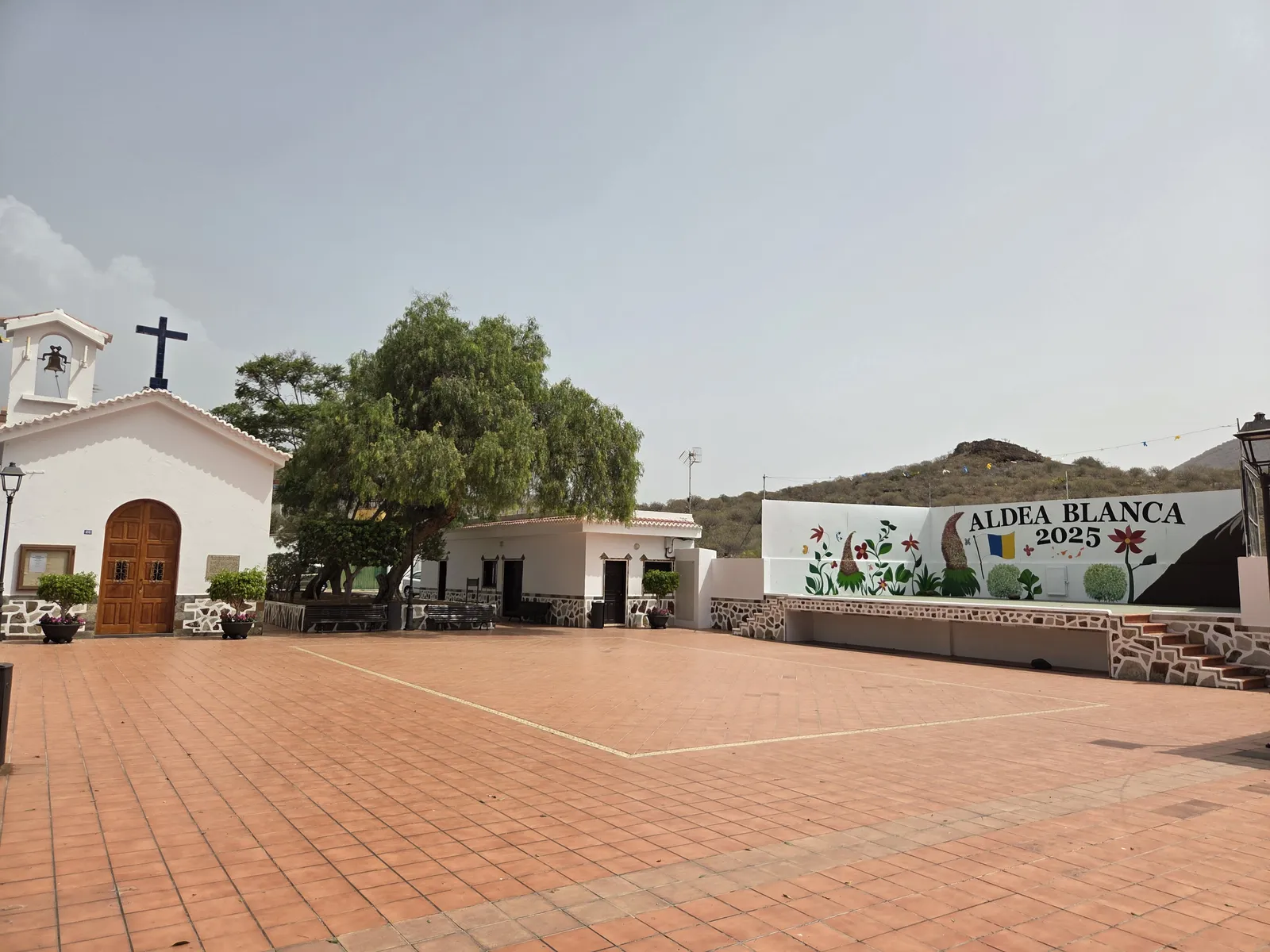 Terracotta-paved square in Aldea Blanca beside a white chapel, trees and a village mural
