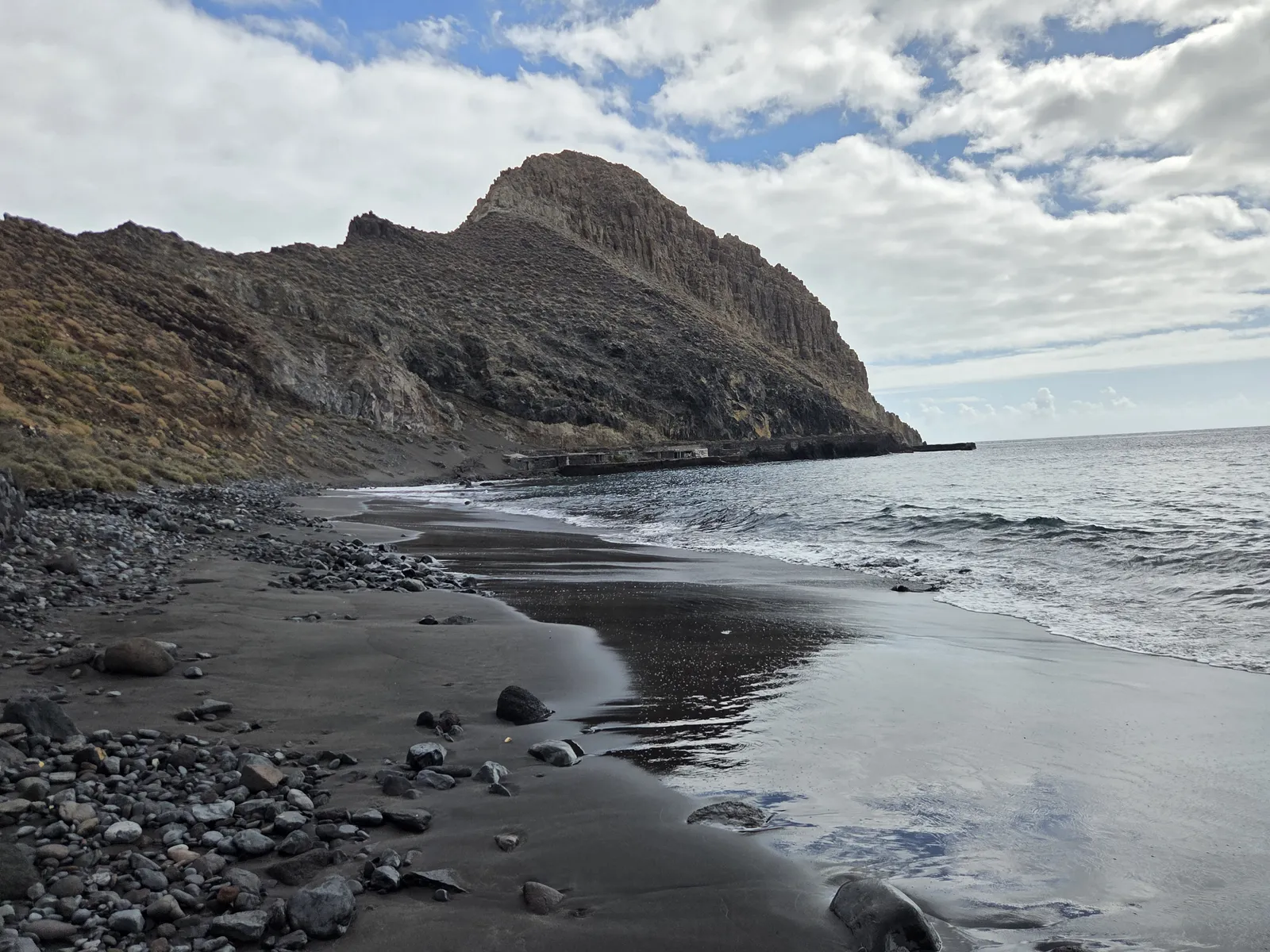 Dark wet sand, rounded stones and breaking waves beneath Antequera’s rocky headland