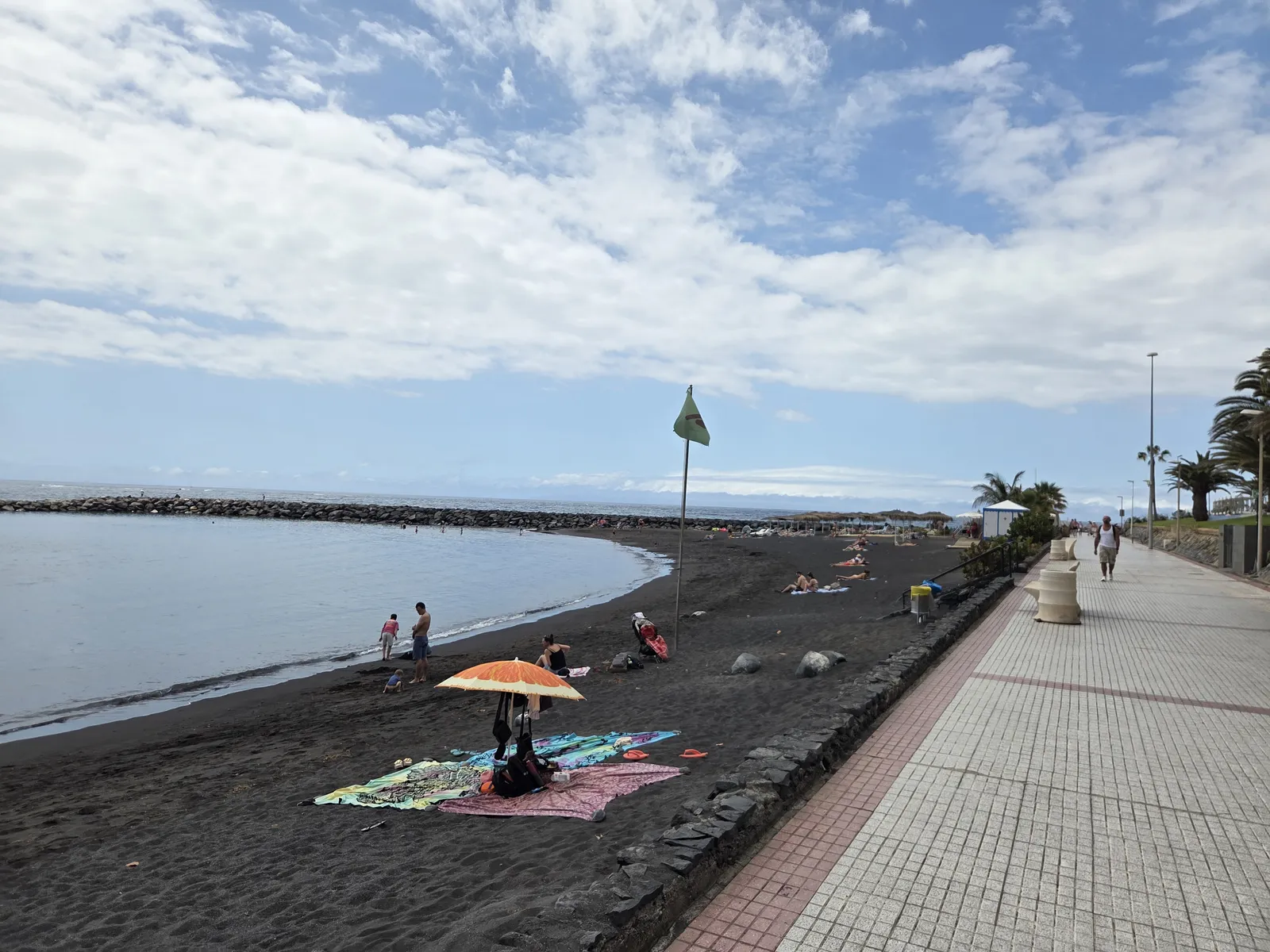Dark sand, sunbathers and a paved promenade beside a breakwater in the El Duque beach area
