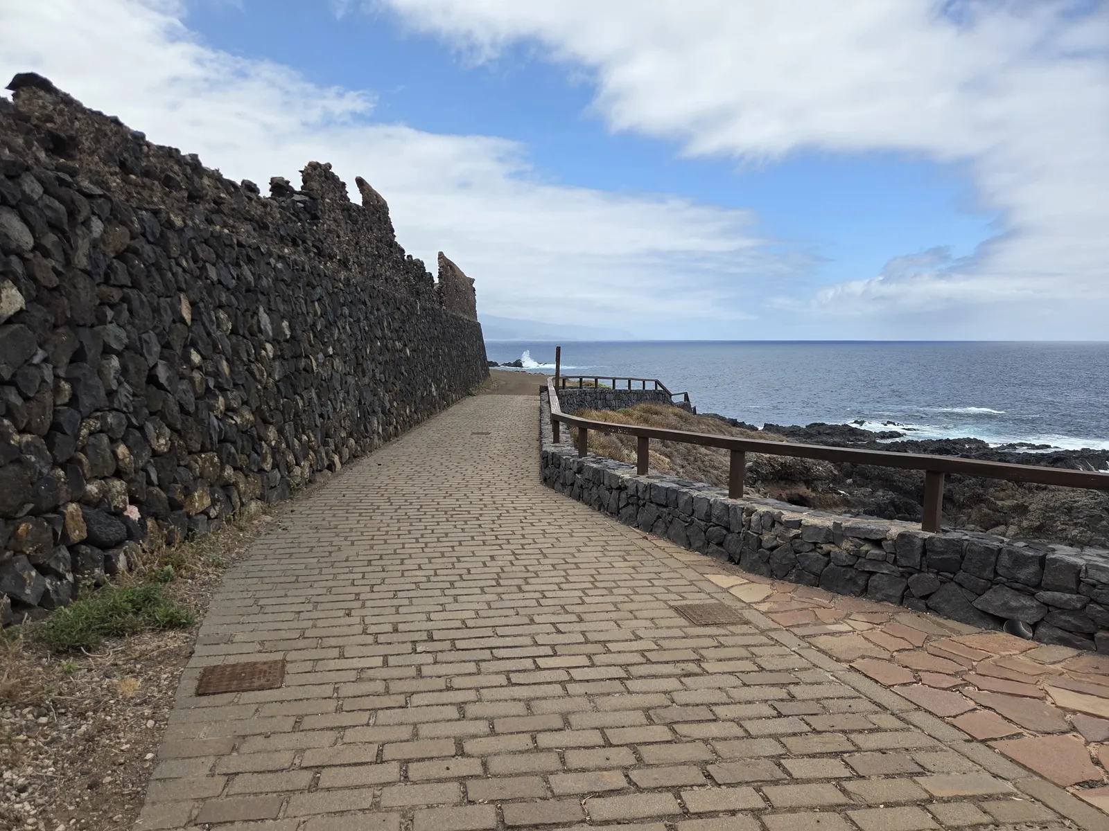 Paved coastal path beside a tall volcanic stone wall and wooden railing in El Sauzal