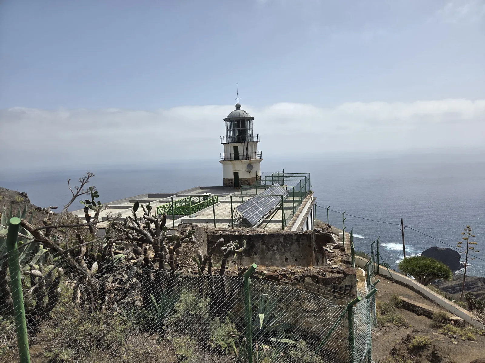 Faro de Anaga’s white lighthouse tower above fenced buildings and solar panels overlooking the Atlantic