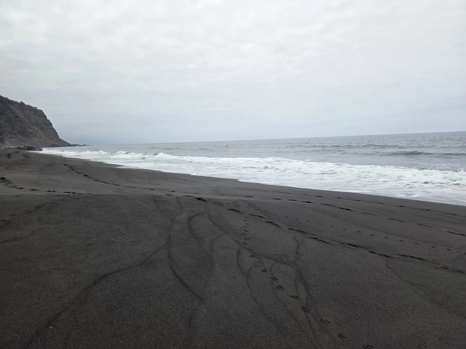 Broad stretch of black volcanic sand at Garañona Beach with breaking waves beneath an overcast sky