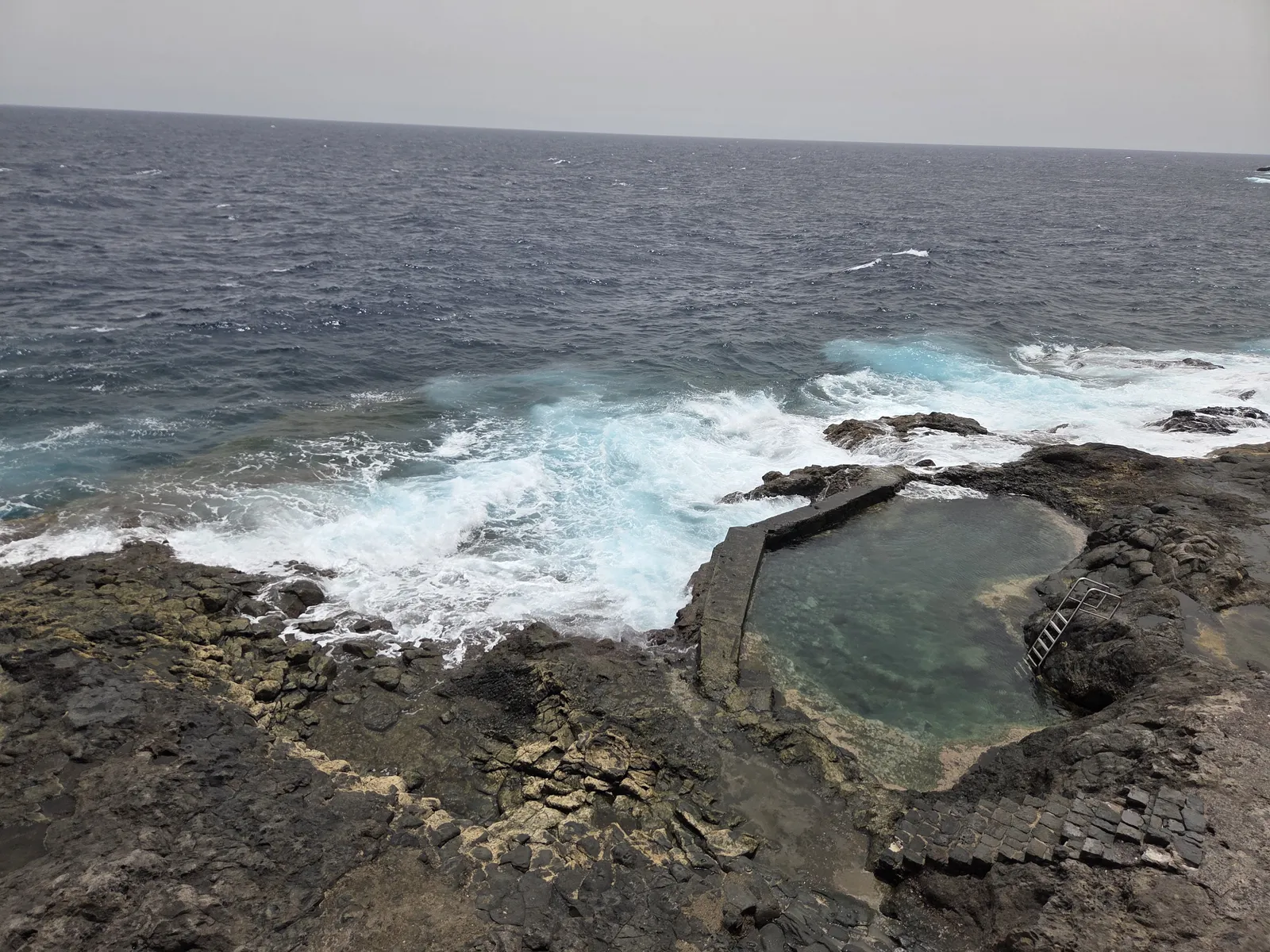 Small walled seawater pool among volcanic rocks at La Caleta, with waves breaking beyond it