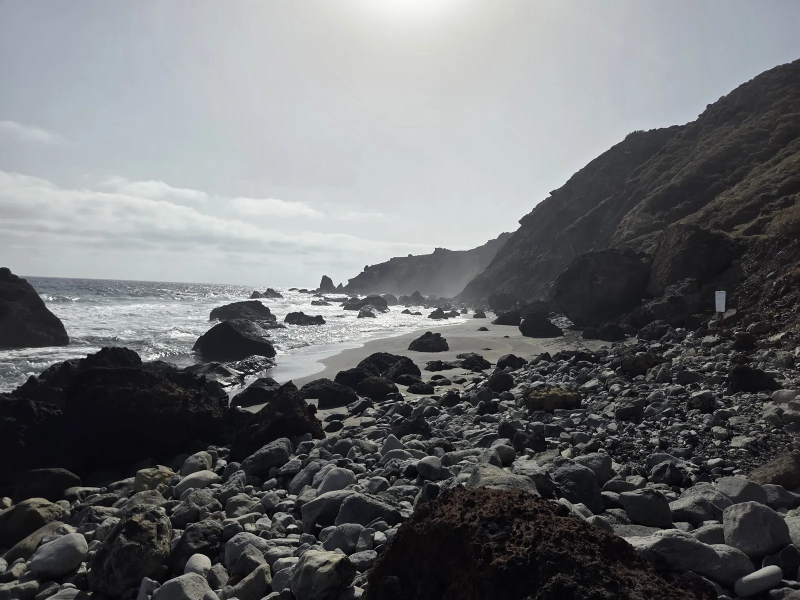 Rounded stones and large dark boulders beside a narrow strip of sand beneath the slopes at Las Breñas
