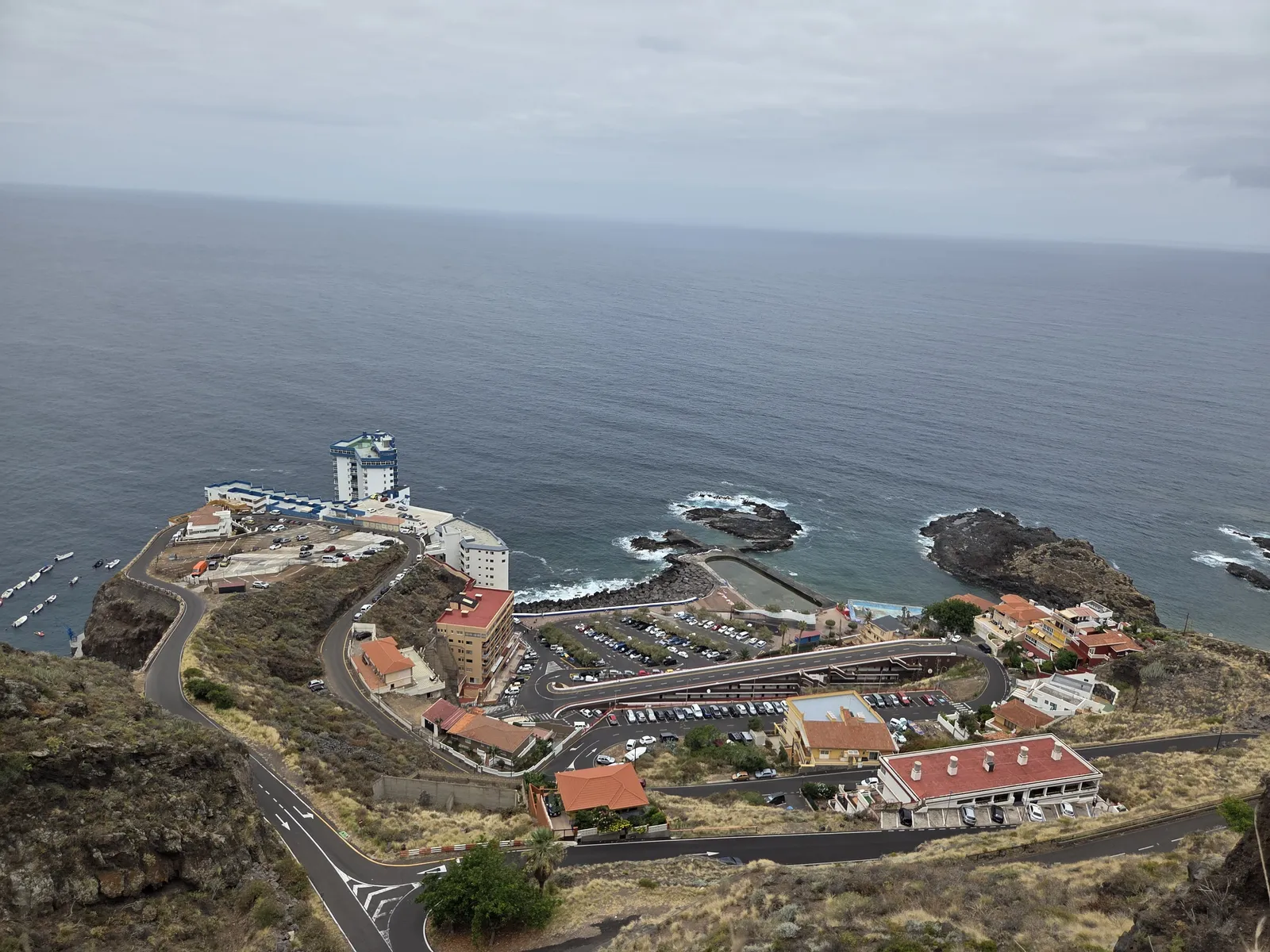 Winding access road descending to Mesa del Mar, with waterfront buildings, parking and a seawater pool