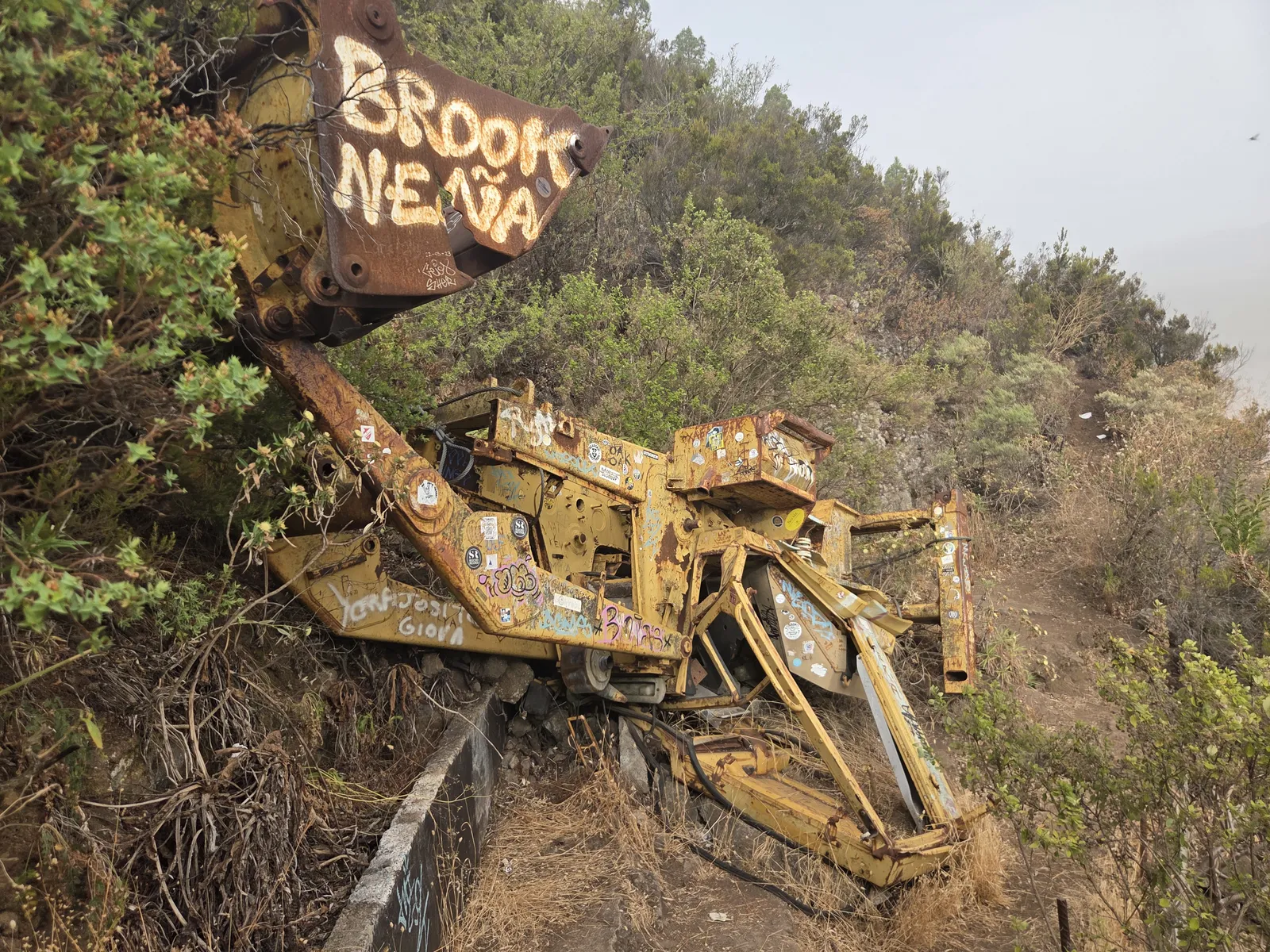 Wrecked yellow excavator lodged against a scrub-covered slope beside Mil Ventanas