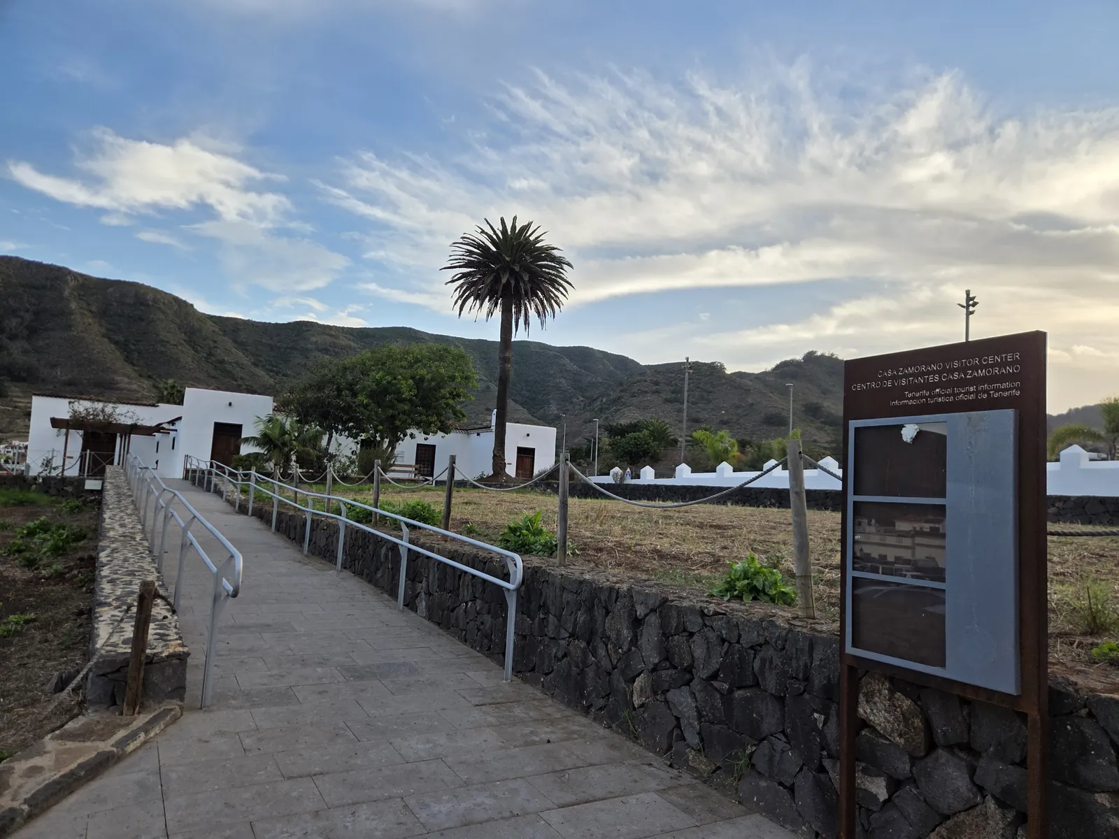 Paved approach to Los Zamorano beside stone walls, cultivated ground and a tall palm