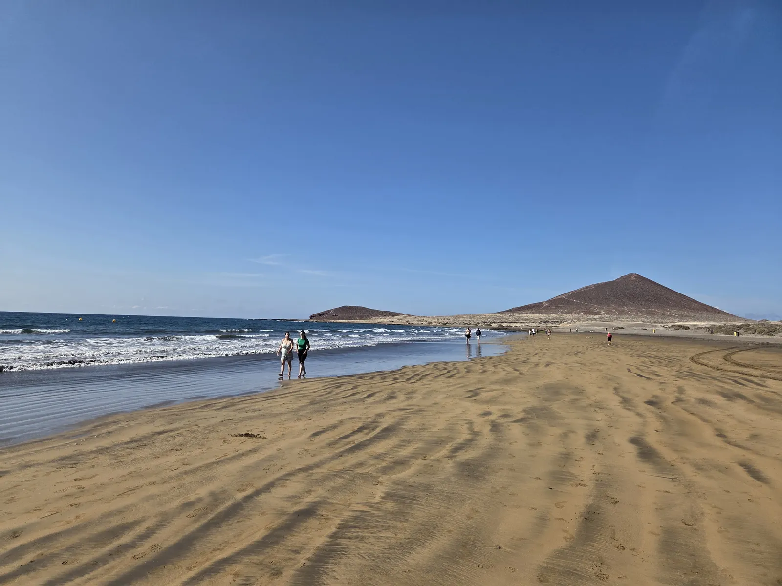 Walkers beside the sea on El Médano beach with Montaña Roja beyond
